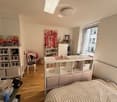Cozy bedroom with a bed, a white table with two glasses, a chair, a shelf with storage boxes, and various decorative items including artworks and a vase.