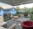 Balcony with a sofa, chairs, a small table, and flower pots, overlooking a residential area with houses and trees.