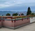 Roof terrace with red metal railing, tiled floor, and scenic city and mountain view.