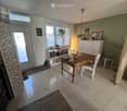 Bright room with tiled floor, large windows, a wooden table with chairs, a white cabinet, a mirror, and a shelf with books and decorative items.