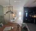 A modern, open-concept living space with a wooden dining table, laptop, pendant light, and a visible bedroom area with a bed and pillows. The kitchen features dark cabinets and an oven.