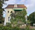 Two-story house with a balcony, surrounded by greenery and trees.