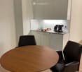 A modern kitchen area with a round wooden table, two black office chairs, a coffee machine, a sink with a faucet, and light-colored cabinets.