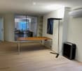 A modern, minimalist room with a wooden floor, a large wooden table, a chair, a floor lamp, a shelf, an air conditioner, and a fan.