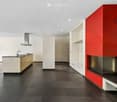 Modern kitchen with dark tiled flooring, white and beige cabinets, a central island with a sink and stove, and a built-in fireplace with a red accent wall.