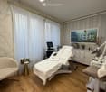A well-lit, modern spa treatment room with wooden flooring, a white massage table, a small round table with a chair, a screen, a cabinet, and a mirror.