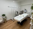 A well-organized massage or treatment room with a massage table, a small round table, a chair, a fan, a towel warmer, a screen, and plants.