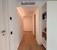 A modern hallway with wooden flooring, white walls, built-in shelving on one side, and recessed lighting. The hallway features a closed door at the end and a built-in cabinet.