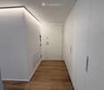 Modern hallway with wooden flooring, white walls, and built-in lighting along the ceiling. Features a closed door at the end and recessed wall panels.