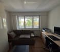 A cozy living room with a sofa, desk, chair, screen, printer, and a lamp. The room has large windows with curtains and a view of greenery outside.