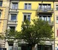 A multi-story residential building with yellow and grey facades, ornate balconies, and large windows. The ground floor features commercial storefronts with green doors and glass windows.