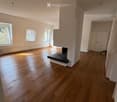 Empty room with wooden flooring, white walls, and a fireplace. Three windows allow natural light to enter, and the space is completely unfurnished.