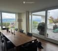 Dining room with a large wooden table, black chairs, a vase with white flowers, and a floor lamp. Large windows offer a scenic view of the landscape, with a small balcony area visible through the glass.