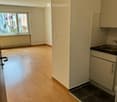 Empty room with light wooden flooring, white walls, and a small kitchen area featuring a sink and cabinets. A large window offers a view of the street.