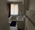 Modern bathroom with a sink, toilet, bathtub, radiator, and a small stool near the sink. The room features a window with sheer curtains and a dark bathmat on the floor.