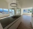 Modern kitchen with light wooden flooring, sleek grey cabinets, and a dark granite countertop. Features large windows with scenic mountain views and a round pendant light fixture.