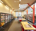 Bright, modern library or study area with large windows, bookshelves, tables, chairs, a computer, and a chessboard on a table.