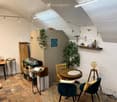 Cozy attic room with exposed wooden beams, brick floor, round wooden table with two chairs, small round stool with lamp, shelves with plants and decorative items, and a woven rug.