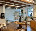 A cozy, well-lit room with wooden floors and exposed wooden beams. The space features a round wooden table with various decorative items, a small stool, a cabinet with drawers, a fan, a mirror, and several plants. The room also includes a glass display ca