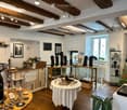 A well-lit boutique interior with exposed wooden beams, white walls, and wooden flooring. Shelves and display cases hold various decorative items, including vases, necklaces, and artworks. A central round table with a white tablecloth displays additional