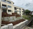 Modern multi-story building with large windows and balconies, surrounded by landscaped garden with stone walls and small plants.