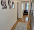 A well-lit hallway with wooden flooring, a patterned rug, and three framed black-and-white pictures on the wall. The hallway leads to a living room area with a sofa and side table visible in the background.