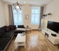Modern living room with wooden flooring, large windows, grey sectional sofa, white coffee table, TV stand, side table, wall shelves, and a geometric hanging light fixture.