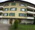 A three-story residential building with yellow walls, green shutters, and multiple balconies. Features include a garage, a small garden with a bench, and a driveway.