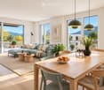 Modern living and dining area with large windows offering a scenic mountain view. Features a green sectional sofa, wooden coffee tables, a dining table with chairs, and decorative items including a vase with candles and a bowl of fruit.