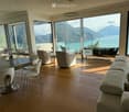 Modern living room with large windows offering a scenic mountain and lake view. Features a sectional sofa, coffee table, dining table with chairs, and several lamps.