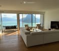 Modern living room with large windows offering a scenic mountain and lake view, featuring a grey sectional sofa, coffee table, armchairs, ottoman, and a television.