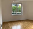 Empty room with light wooden flooring and a large window with white frame, showing greenery outside.