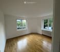 Empty room with wooden flooring, white walls, and two windows offering a view of greenery outside.