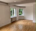 Empty room with wooden flooring, white walls, and two windows showing greenery outside. Radiator under one window.