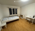 A small, tidy bedroom with a single bed, a desk with a chair, and a small storage unit. The room features light wooden flooring and white walls, with a window providing natural light.