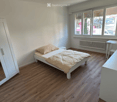 Cozy bedroom with wooden flooring, a single bed with a patterned blanket, a white wardrobe with a mirror, and a small table near the window.