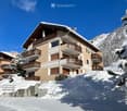 A multi-story residential building with balconies, surrounded by snow-covered trees and mountains under a clear blue sky.