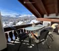 Balcony with a round glass table, two chairs, one with a fur throw, overlooking a snowy mountain landscape.