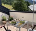 Balcony with a table set for two, featuring wine glasses, plates of salad, and flower pots, overlooking a scenic mountain landscape.