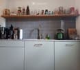 Modern kitchen with grey cabinets, marble countertops, and grey subway tiles. Coffee maker, teapot, cutting board, and various kitchen utensils are visible on the counter. Shelves above hold jars, cups, and other kitchen items.