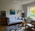 Modern living room with a light grey sofa, wooden chair, round wooden stool, side table, floor lamp, picture frames, books, and plants near the window.