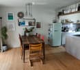 Bright kitchen and dining area with wooden floors, a light blue refrigerator, a wooden dining table with chairs, and open shelving with various items. The space features framed artwork, a potted plant, and a hanging light fixture.