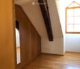Empty attic room with wooden floor, built-in wooden cabinets, exposed wooden beam, and a single window letting in natural light.
