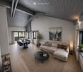 Modern living and dining area with exposed wooden beams, light grey sofa, coffee table, dining table with chairs, and decorative plants.