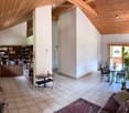Spacious living and dining area with wooden beams, tiled floor, and large windows. Features a sofa, dining table with chairs, bookshelf, and plants.