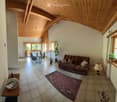 Bright living room with exposed wooden beams, tiled floor, and large windows. Features a brown sofa with decorative pillows, a dining table with chairs, a patterned rug, and a side table with a lamp.