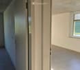 Empty room with tiled floor, white walls, and two windows with blinds. One door is open, revealing a view of greenery outside.