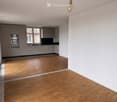 Empty room with light wooden flooring, white walls, and a small kitchen area with cabinets and a sink visible in the background.