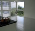 Bright, minimalist room with large windows, white tiled floor, and a small black countertop holding a bowl of fruit. A white vase with dried branches stands near the windows.