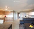 Modern open-plan living and dining area with a sleek black sectional sofa, a wooden coffee table with fruit, a dining table with chairs, and a contemporary kitchen featuring light wood cabinets, stainless steel appliances, and a central island.